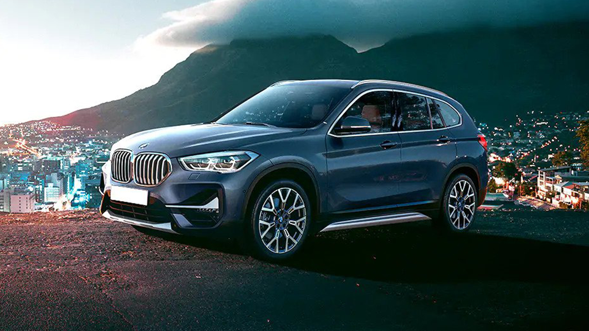 A dark blue BMW X1 is parked on a hill overlooking a city at dusk.