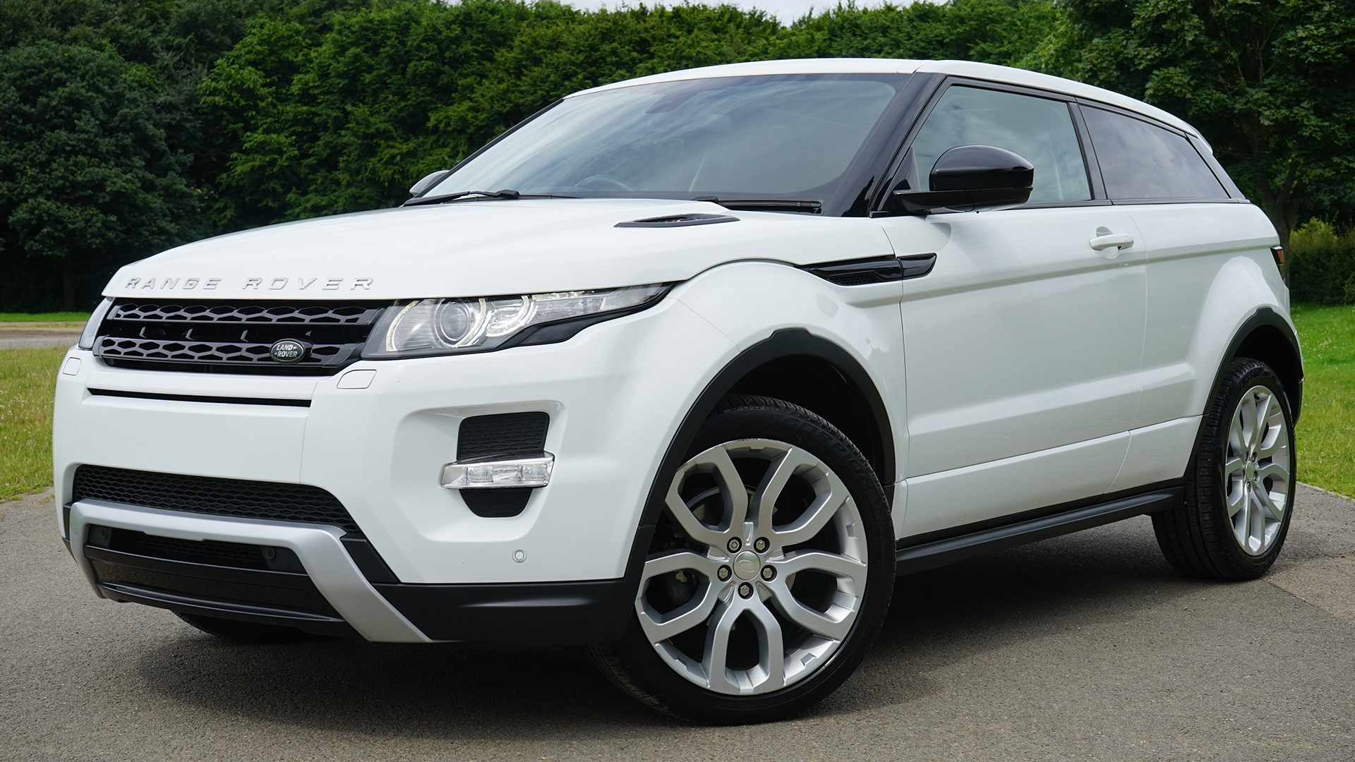 A white Range Rover Evoque is parked on a paved road with trees in the background.